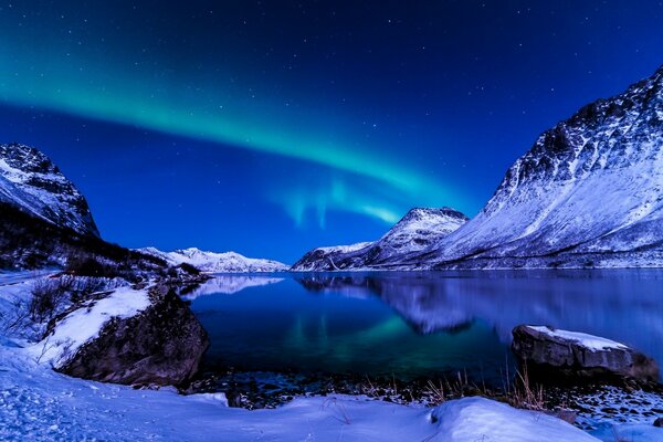 Paisagem ártica de um lago de montanha congelado em margens cobertas de neve sob o céu com luzes do Norte
